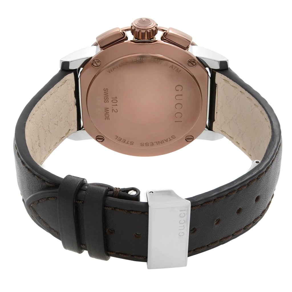 An image of a new Gucci men's wristwatch viewed from the back, showcasing the stainless steel screwback case with the brand name engraved around the perimeter. The watch is positioned at a direct angle with the camera focusing closely on the closure, highlighting the deployant clasp on a black leather band. The inside of the strap is visible, revealing a beige color with texturing, and the watch is oriented such that the band forms a semi-circle at the bottom of the frame with the clasp slightly opened to the right side. A protective sticker is attached to the band, indicating the item's new condition with defects.