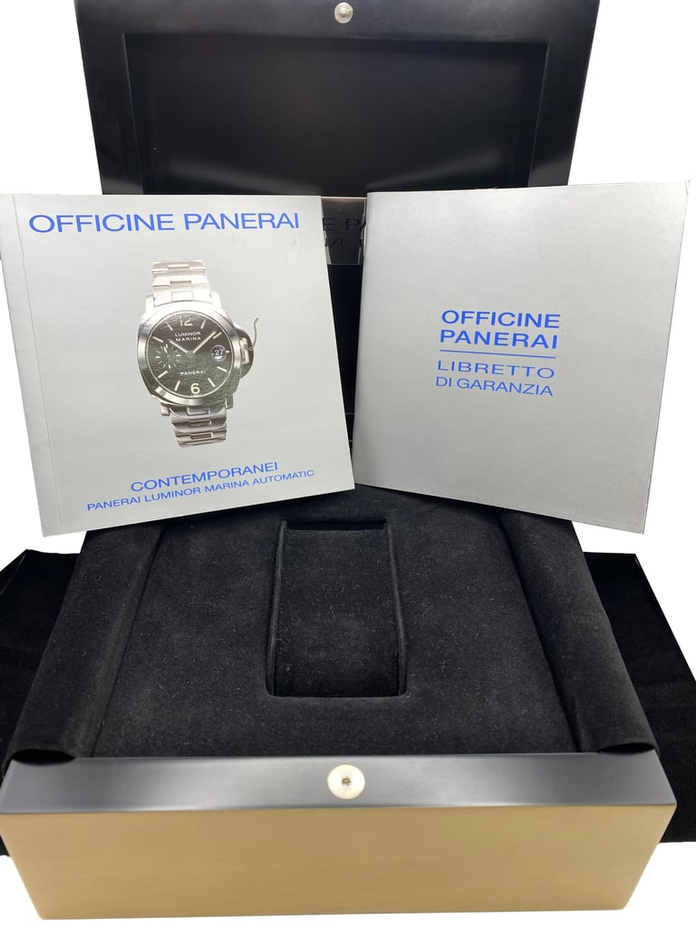 An image of a pre-owned Officine Panerai men's wristwatch packaging, viewed from a slightly elevated angle. The watch itself is not present. In the foreground, a black velvet-lined box with an empty watch cushion is open. Behind it, two light blue documents stand upright: to the left is a booklet with 'OFFICINE PANERAI CONTEMPORANEI PANERAI LUMINOR MARINA AUTOMATIC' and to the right is a warranty booklet with 'OFFICINE PANERAI LIBRETTO DI GARANZIA.' The background features a portion of the black exterior box. The focus is centered on the documents and the interior watch box, providing a full view of the watch's packaging and accompanying materials.