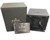 An image of an unworn Edox men's wristwatch packaging, displaying the brand's box and documents without the watch itself. The textured gray box, bearing the Edox logo and the words "Maître Horloger Les Genevez depuis 1884," is positioned on the left and slightly angled to face the camera. An Edox International Warranty card is visible, partially covered in protective plastic wrapping. The items are arranged closely together, shot from a high angle in medium close-up, capturing the full view of the items meant to accompany the wristwatch.