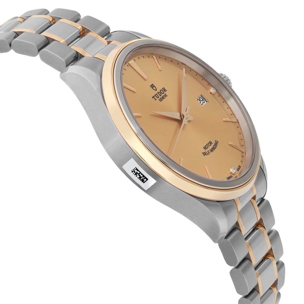 An image of a wristwatch shown from a side profile view, opposite the crown. The watch is unworn and features a dual-tone bracelet with a metallic finish. The face of the watch is angled slightly upward, showcasing its gold-colored dial and date function. The image is taken from a close distance, highlighting the sleek design and craftsmanship.