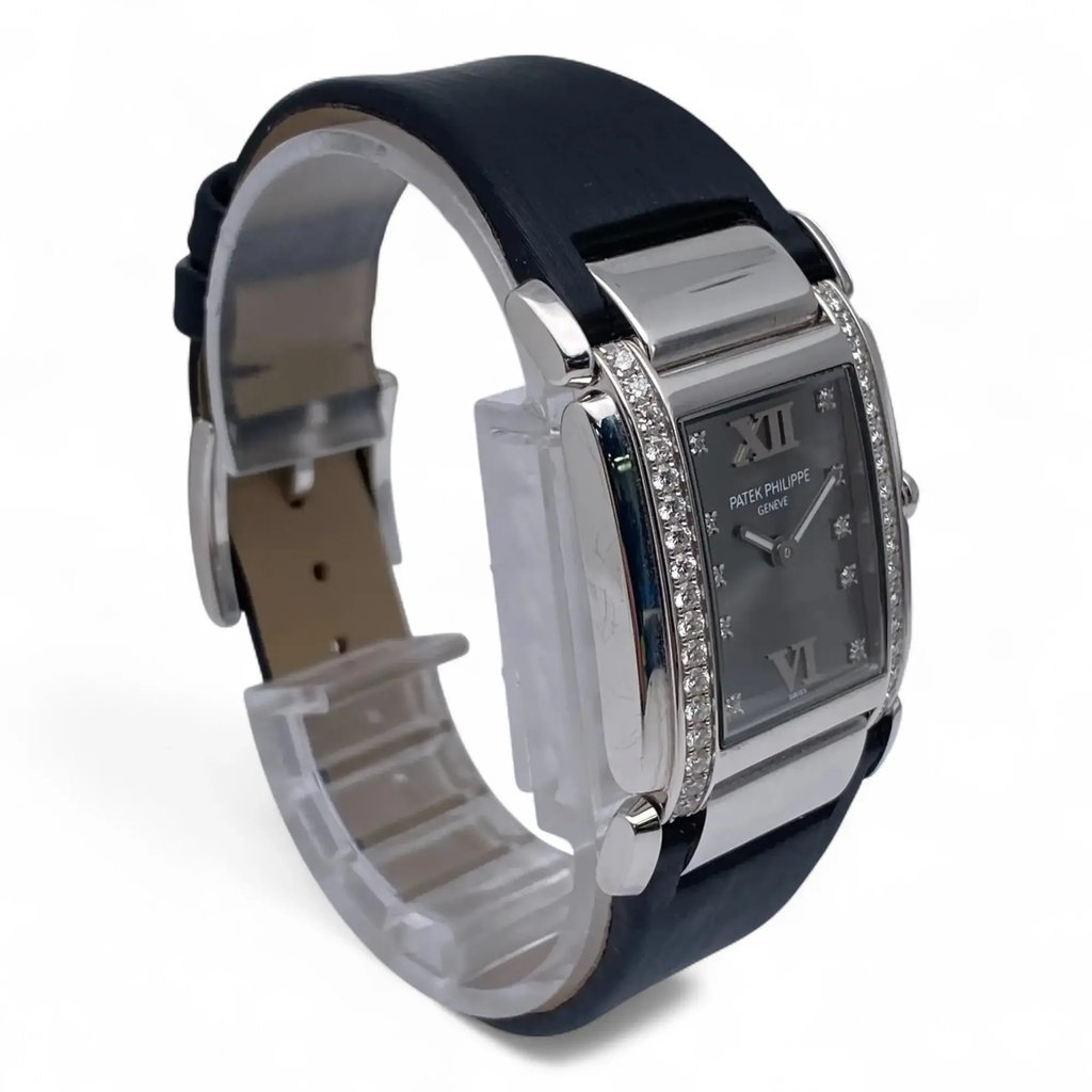 An image of a pre-owned Patek Philippe women's wristwatch. The watch is positioned at a slight angle, facing left, showcasing its elegant rectangular face adorned with diamond accents. The dark strap is visible, and the watch is captured from a medium distance, highlighting its luxurious details and craftsmanship.