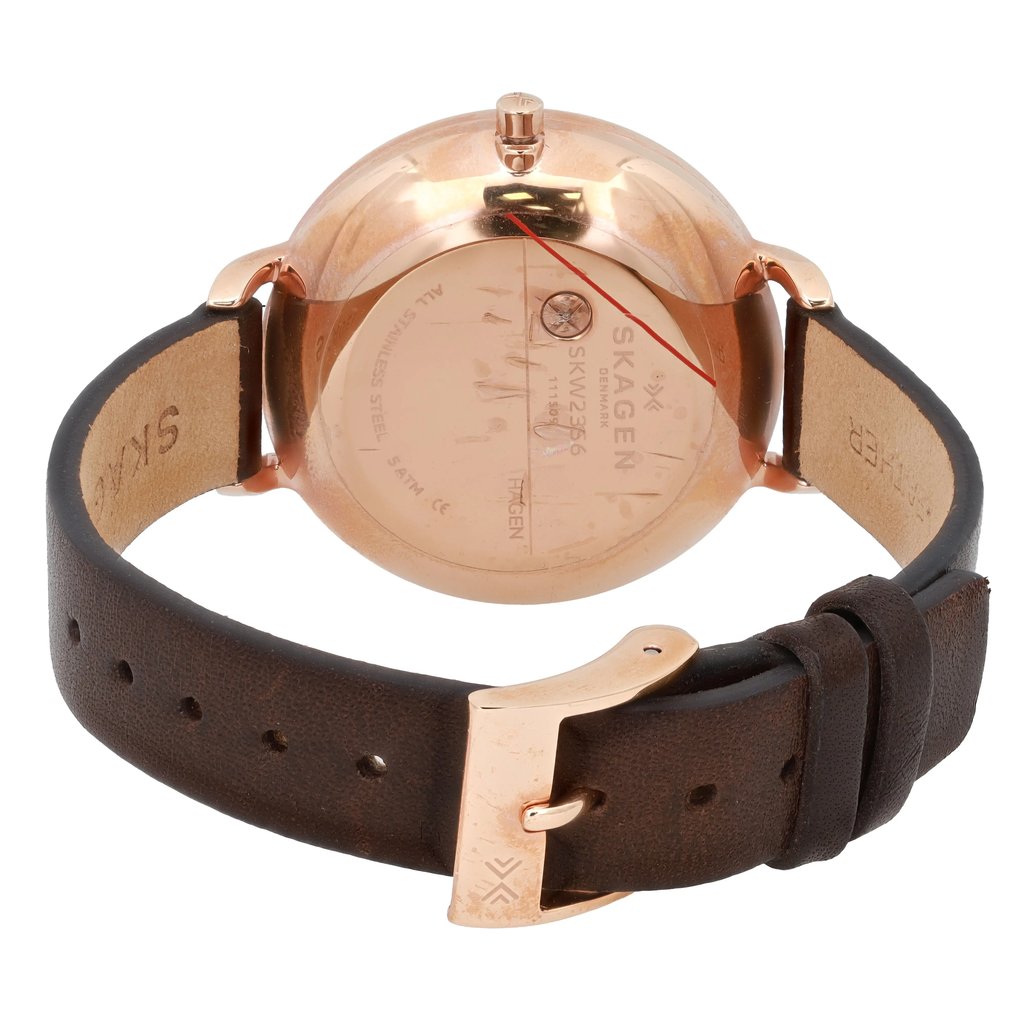 An image of a pre-owned Skagen wristwatch positioned at a slight angle, viewed from the back. The focus is on the rose gold case and the embossed brand details on the back cover. The dark brown leather strap is visible, with the buckle prominently featured in the foreground. The watch is captured from a close distance, highlighting the texture and details of the materials.