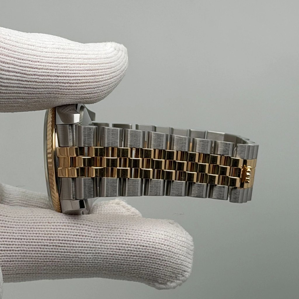 An image of a pre-owned Rolex wristwatch held horizontally by a gloved hand, viewed from the side. The focus is on the bracelet, which features a two-tone design with gold and silver links. The watch case and crown are partially visible, with the fluted bezel adding a touch of elegance. The angle captures the watch's intricate details, emphasizing its craftsmanship. The photo is taken at a close distance, highlighting the texture and finish of the materials.