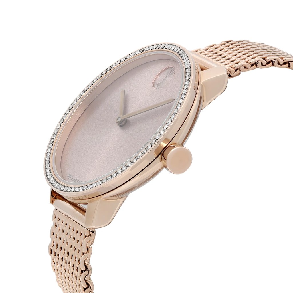 An image of a pre-owned wristwatch taken from a side profile view, showcasing the crown prominently. The watch has a rose gold case with a minimalist pink dial. The bezel is adorned with small crystals, adding an elegant touch. The mesh band is also rose gold, complementing the overall design. The angle highlights the watch's sleek and refined appearance.