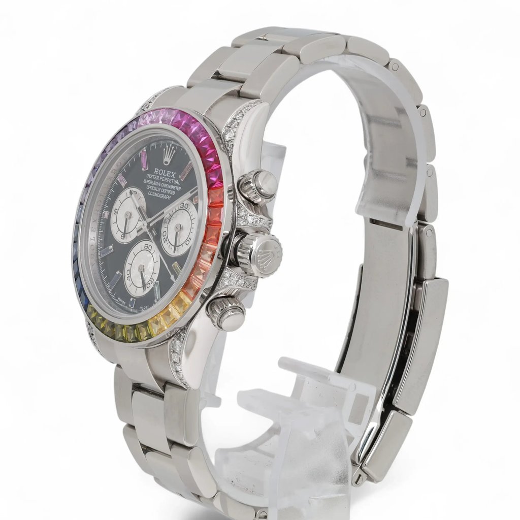 An image of a pre-owned men's Rolex wristwatch, angled slightly from the front and left side, showcasing its stainless steel bracelet and case. The watch features a multicolored bezel, chronograph functionality, luminous hands, and small seconds subdial. The watch is positioned upright on a transparent stand with a close-up view highlighting its intricate details.
