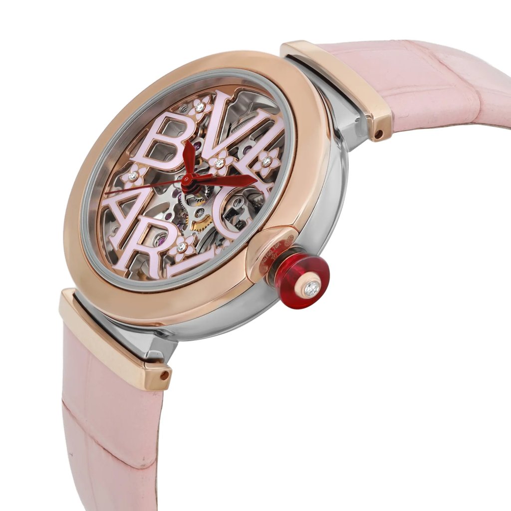 An image of a pre-owned wristwatch captured from a side profile view, showcasing the crown prominently. The watch features a pink strap and a round face with intricate internal mechanics visible through the transparent dial. The brand's letters are prominently displayed across the face. The crown is accented in red and set with a small gem. The angle highlights the watch's modern and stylish design.