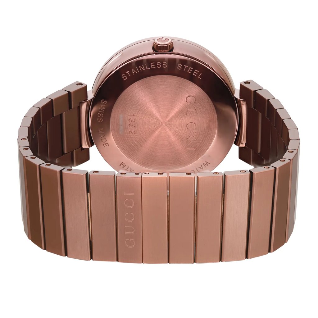 An image of a men's Gucci wristwatch, display model, viewed from the back showing the caseback and closed clasp. The watch is photographed at a slight angle with the caseback centered and the stainless steel band extending to the left and right, partially out of frame. The snap caseback is engraved with the Gucci brand and other details, and the hidden fold clasp is visible but in the closed position. The round case and band material both appear to be a uniform rose gold tone. The image presents the watch in close-up, capturing fine details such as minor blemishes due to storage.