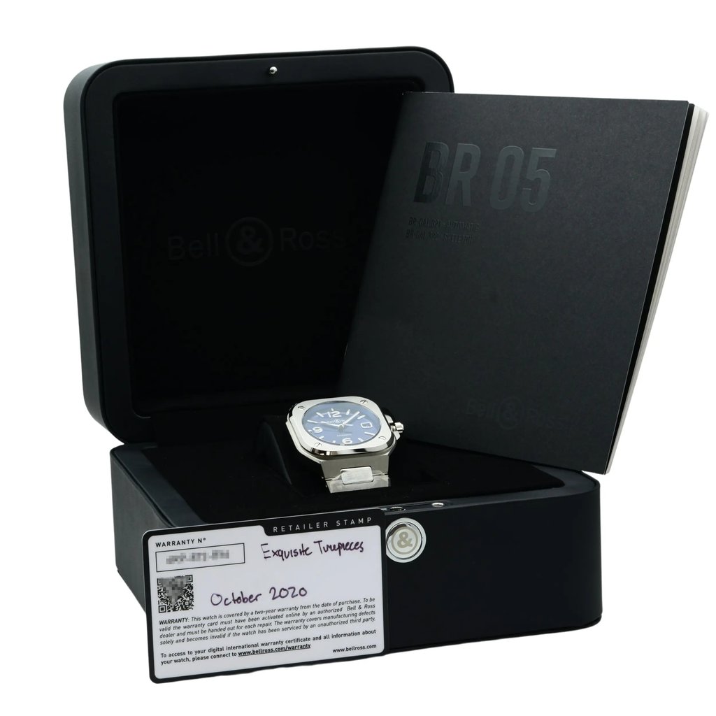 An image of a Bell & Ross men's wristwatch with its box and documents. The watch is positioned in the foreground at a slight right angle, resting on its side on top of the black box with the Bell & Ross logo visible inside the lid. The box is partially open, angled towards the camera, and a warranty card with a visible date of "October 2020" is placed on the front of the box. In the background, slightly out of focus, is a black booklet with "BR 05" embossed on the cover. The overall view is from a medium distance, presenting the entire set-up clearly.