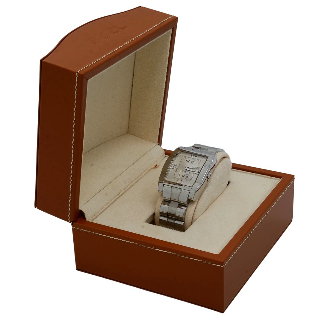 An image of a pre-owned wristwatch positioned at a slight angle, displayed inside an open, brown leather box with beige interior. The watch has a metallic band and is placed on a cushioned holder, positioned slightly towards the right side of the box. The shot is taken from a close distance, capturing the full view of the box and watch.