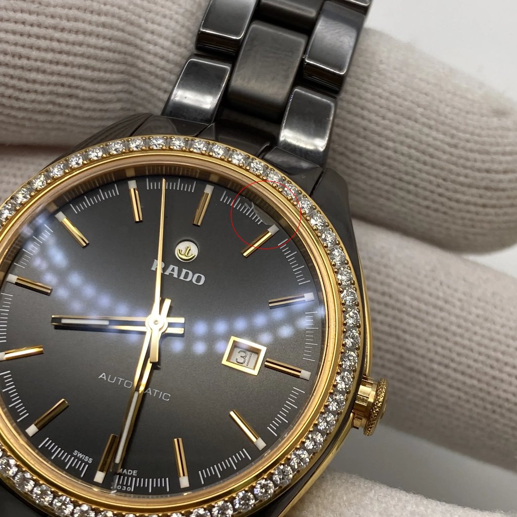 An image of a pre-owned women's luxury wristwatch by Rado. The photo shows a front close-up view of the watch face, highlighting its round shape. The dial is brown with non-numeric, stick indexes, and features a gold bezel adorned with small stones. The hands are luminous, and there is a date indicator at the three o'clock position. The band is made of ceramic, and the case combines ceramic and stainless steel materials. The watch is held by a gloved hand, emphasizing its good condition. The watch has tiny scratches on the gold-tone surface. The dial has damage at the 1 o'clock position.

