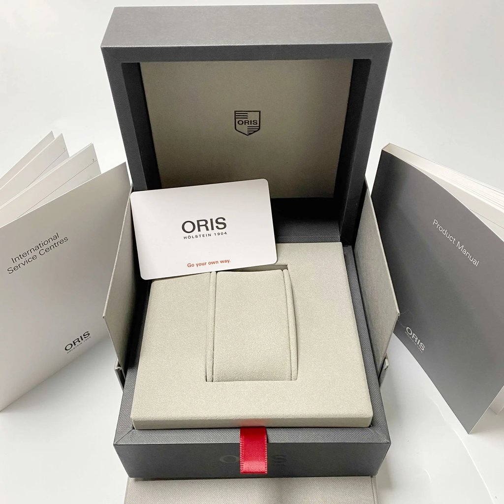 An image of an Oris wristwatch display box and accompanying documents. The box is open, showcasing a padded watch holder inside. Positioned centrally is a branded card with the Oris logo. Surrounding the box are various manuals, including an International Service Centers booklet on the left and a Product Manual on the right. The photo is taken from a slightly elevated angle, capturing the full view of the box and documents at a close distance.