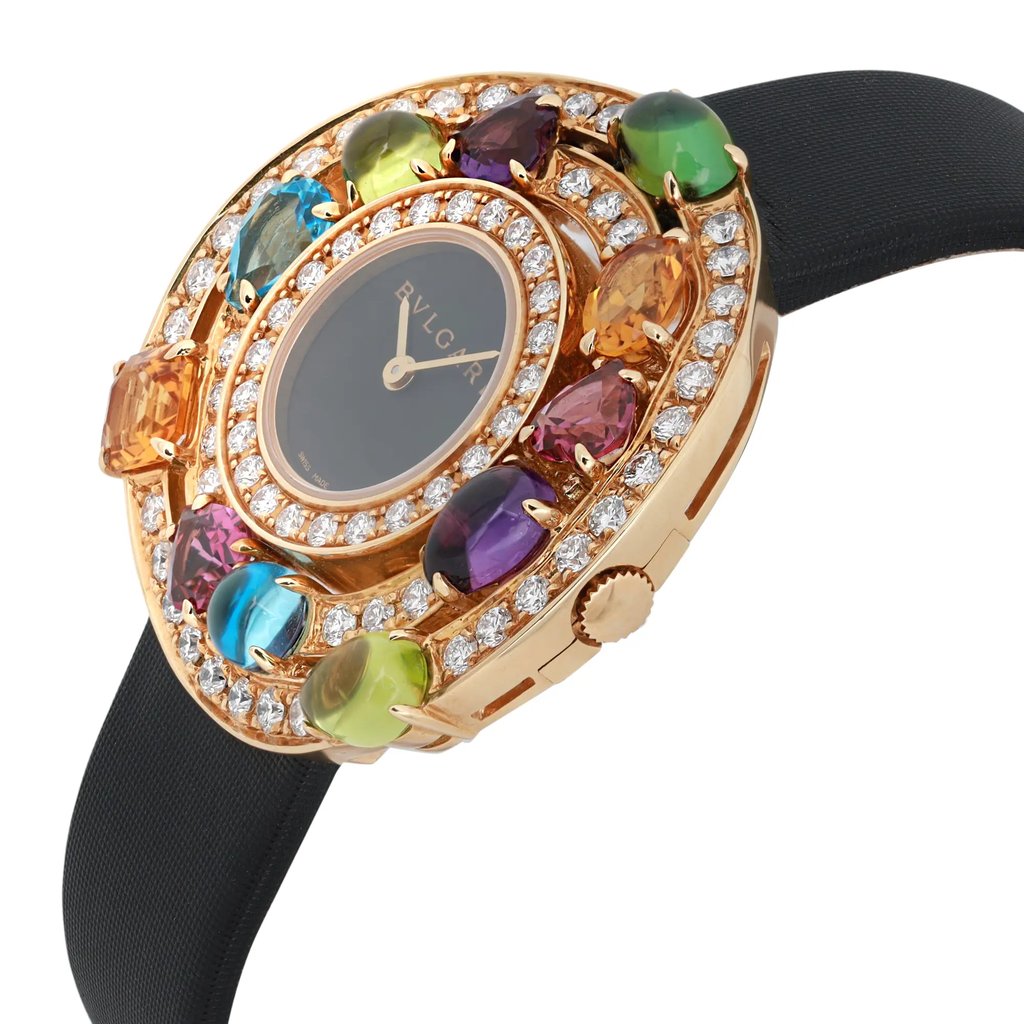 An image of a pre-owned women's Bvlgari wristwatch viewed at an angle, showcasing its opulent design. The watch has a black face with gold hands and is encircled by an array of colorful gemstones and diamonds set in a gold bezel. The watch strap is black and positioned in the background. The image is taken from a close-up perspective, highlighting the intricate details and luxurious craftsmanship.