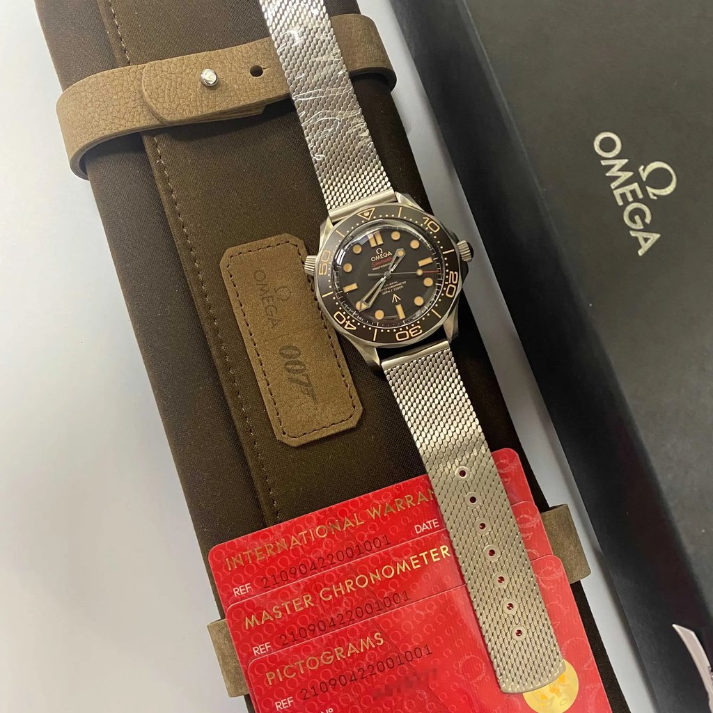 An image of a men's OMEGA wristwatch, displayed as a model with its box and documents in full view. The watch is positioned diagonally across the frame with the top left corner closer to the viewer, showcasing a brown leather strap on the left and a silver metal mesh bracelet on the right. The watch face is visible with a black dial and luminous markers. Below the watch, red warranty and pictogram cards are partially visible, with the OMEGA logo on a black box in the background to the right. The shot is taken from a close, overhead angle, capturing the details of the watch and accompanying materials.