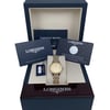 An image of a new wristwatch positioned centrally within an open box, surrounded by documents and warranty cards. The watch features a two-tone metal bracelet and a gold-colored dial. The box lid and documents are angled slightly backward, providing a full view of the contents. The image is taken from a straight-on perspective at a moderate distance, showcasing the brand name on the box and documents.