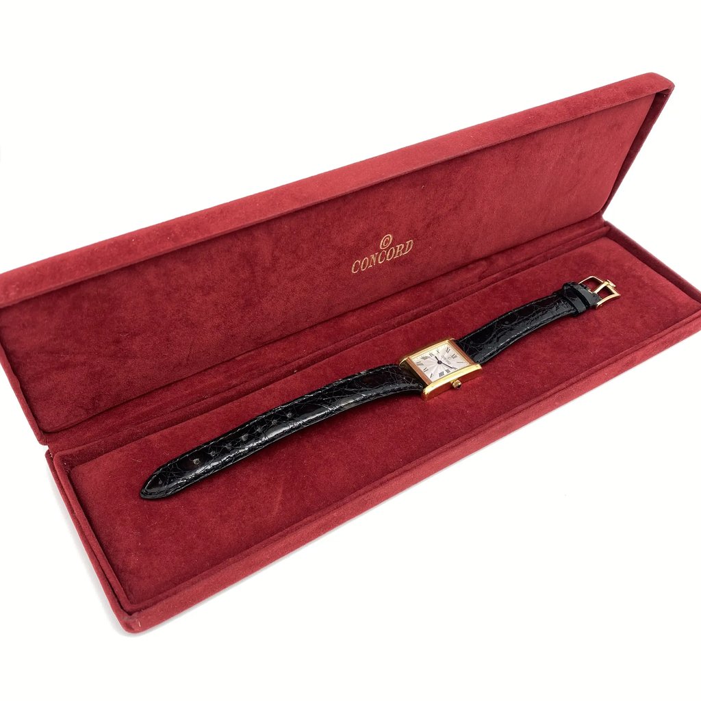 An image of a pre-owned wristwatch positioned inside an open, red velvet box. The watch has a square face and a black leather strap. The box is viewed from a slightly elevated angle, showing the interior lining and the brand name "Concord" on the lid. The photo is taken from a medium distance, capturing the full view of the watch, box, and surrounding documents.
