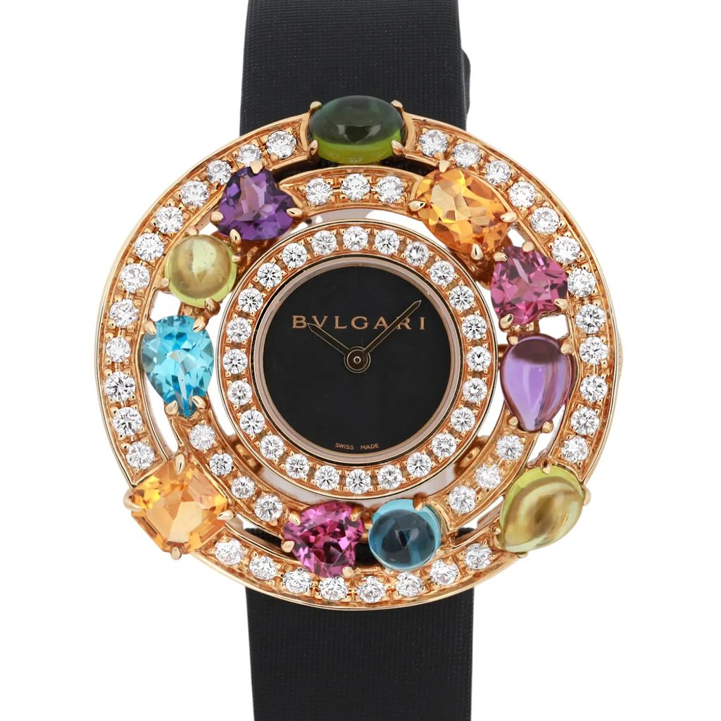 An image of a pre-owned Bvlgari women's wristwatch. The watch face is centrally positioned and viewed directly from the front. It features a black dial with the Bvlgari logo and is surrounded by a luxurious arrangement of colorful gemstones and two concentric circles of diamonds. The strap is black and partially visible at the top and bottom. The image captures the watch in close-up, showcasing its intricate details and vibrant colors at eye level.