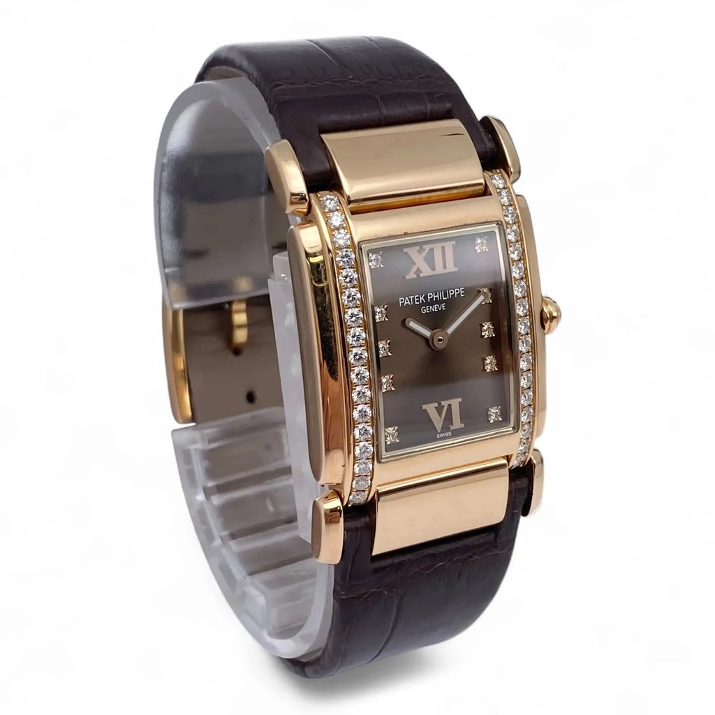 An image of a pre-owned Patek Philippe women's wristwatch positioned at a slight angle from the front, showcasing its elegant rectangular face. The watch features a rose  gold case adorned with diamonds along the sides, a brown leather strap, and a dark dial with Roman numerals at the 12 and 6 o'clock positions. The photograph is taken from a close distance, highlighting the watch's luxurious details.