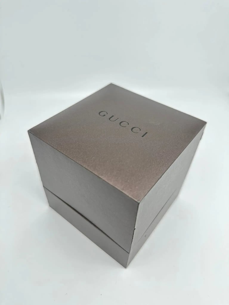 An image of a pre-owned Gucci wristwatch box for men, presented in full view against a white background. The box is captured from a slight high angle, focusing on the top where the "Gucci" logo is prominently displayed in a darker shade, and the box is positioned centrally at a close distance, offering a clear view of its condition and design. The actual wristwatch is not included in the image.