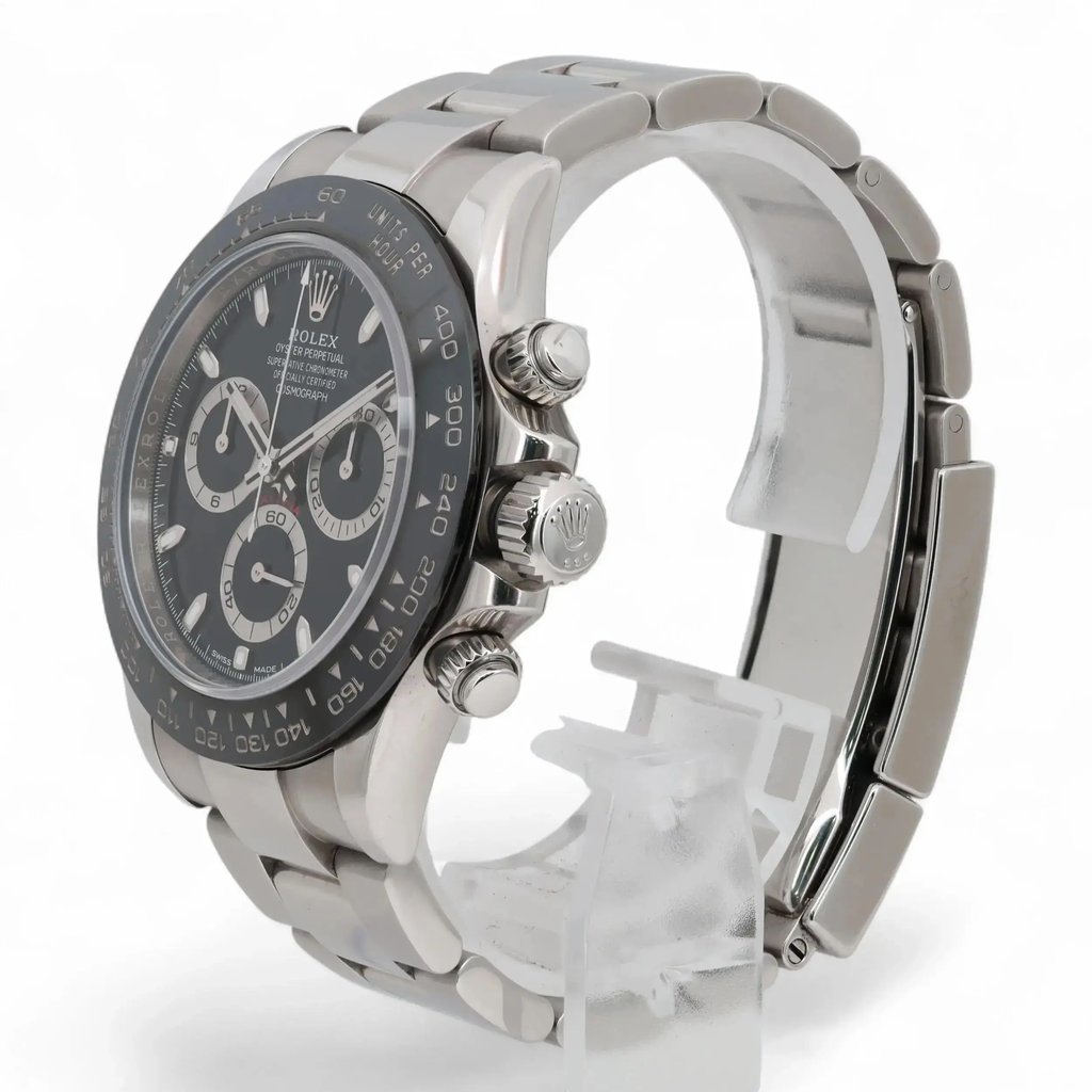 An image of a pre-owned men's Rolex wristwatch, photographed from a slightly elevated angle and positioned to the right. The watch features a stainless steel band and a black dial with chronograph functionality, luminous hands, and a small seconds subdial. The side view showcases the crown and pushers, highlighting the watch's intricate details. The watch is displayed on a transparent stand against a neutral background, captured at a moderate distance.