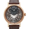 An image of a luxury men's Breguet wristwatch, viewed up close in a wristshot. The watch has a round, rose gold case with a gray dial, featuring intricate mechanical details and a power reserve indicator. The dial lacks hour marks and displays Roman numerals. The leather band is visible at the top and bottom, complementing the elegant design.