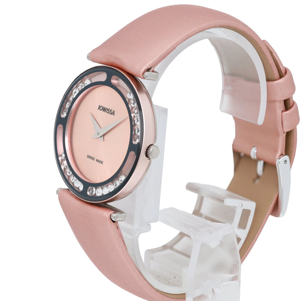 An image of a JOWISSA women's wristwatch positioned at a slight angle, showcasing its round face. The watch has a pink dial with silver hands and is encircled by a decorative bezel with clear stones. The pink strap is partially visible, and the watch is displayed on a clear stand against a white background. The photo is taken from a close distance, highlighting the watch's design and details.