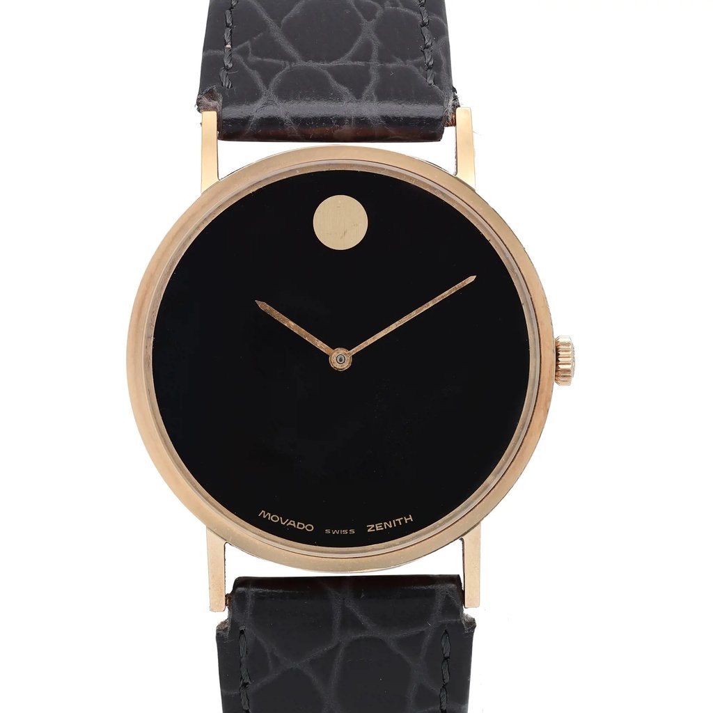 An image of a pre-owned wristwatch taken from a front close-up angle, showcasing the watch face prominently. The watch features a sleek black dial with a minimalistic design, including two gold hands and a gold dot at the 12 o'clock position. The casing is gold-toned, complementing the black leather strap with a textured pattern. The brand names "Movado" and "Zenith" are subtly visible at the bottom of the dial.