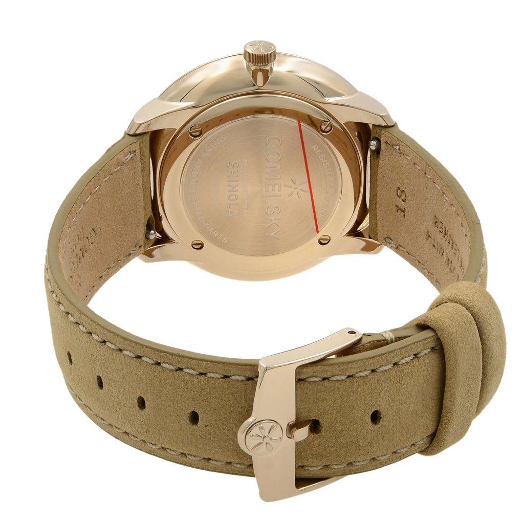 An image of a wristwatch taken from the back view, showcasing the closed clasp. The watch is positioned at a slight angle, highlighting the unworn, pristine condition. The focus is on the gold-toned case back and the beige leather strap, with the brand "GOMELSKY SHINOLA" engraved on the back. The image is captured from a close distance, emphasizing the details of the clasp and stitching on the strap.