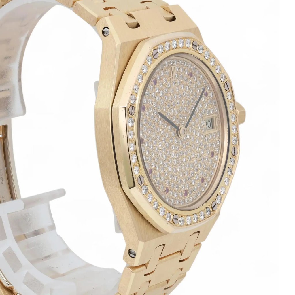 An image of a pre-owned women's Audemars Piguet wristwatch, taken from a slight side angle. The watch features a yellow gold case and bracelet, with a diamond-encrusted bezel. The dial is also set with diamonds and includes a date indicator at the 3 o'clock position. The watch is positioned upright, showcasing its luxurious design, and the image is captured from a close distance to highlight the intricate details.