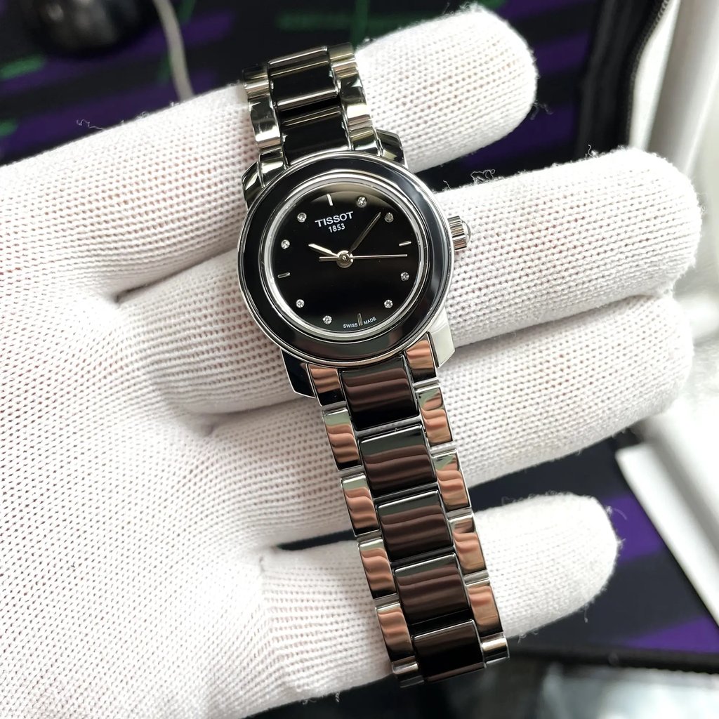 An image of a Wristwatch by Tissot. This Display Model item is designed for Women, featuring a Dress/Formal style. It has a Quartz movement and an Analog display with a Black dial color. The indices include 12-Hour Dial,Diamond Markers,Stick Indexes. The band is made of Ceramic,Stainless Steel and the case is made of Stainless Steel. The watch has a Round shape with a case size of 28 mm and a case thickness of 7 mm.