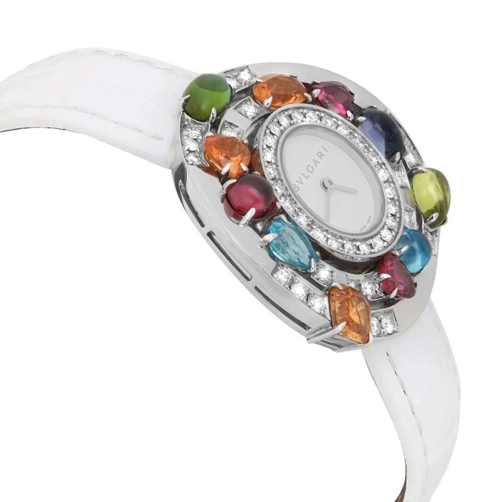 An image of a pre-owned Bvlgari women's wristwatch, taken from a slightly elevated angle showcasing the side and front of the watch. The watch features a white leather strap and a bezel adorned with various colorful gemstones, including green, orange, red, and blue. The face is round, surrounded by a circle of diamonds. The image captures the watch from a close distance, emphasizing the intricate details and vibrant colors.