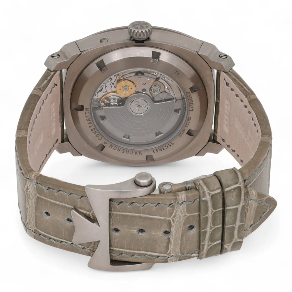 An image of a pre-owned men's Vacheron Constantin wristwatch. The watch is positioned with its back facing the viewer, showcasing the intricate mechanical movement through a transparent case back. The angle is slightly elevated and close-up, highlighting the detailed design of the movement and the engraved brand name around the edge. The leather strap is visible, clasped with a distinctive metal buckle, and presented in a neutral tone.