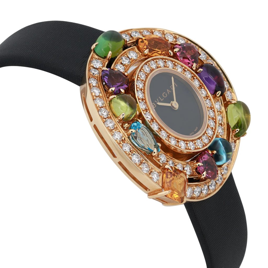 An image of a pre-owned Bvlgari women's wristwatch, captured at a close angle from the side. The watch features a black band and an ornate, circular gold face adorned with colorful gemstones and encircled by sparkling diamonds. The watch face has a dark background with gold hour and minute hands, and the brand name "Bvlgari" is visible.