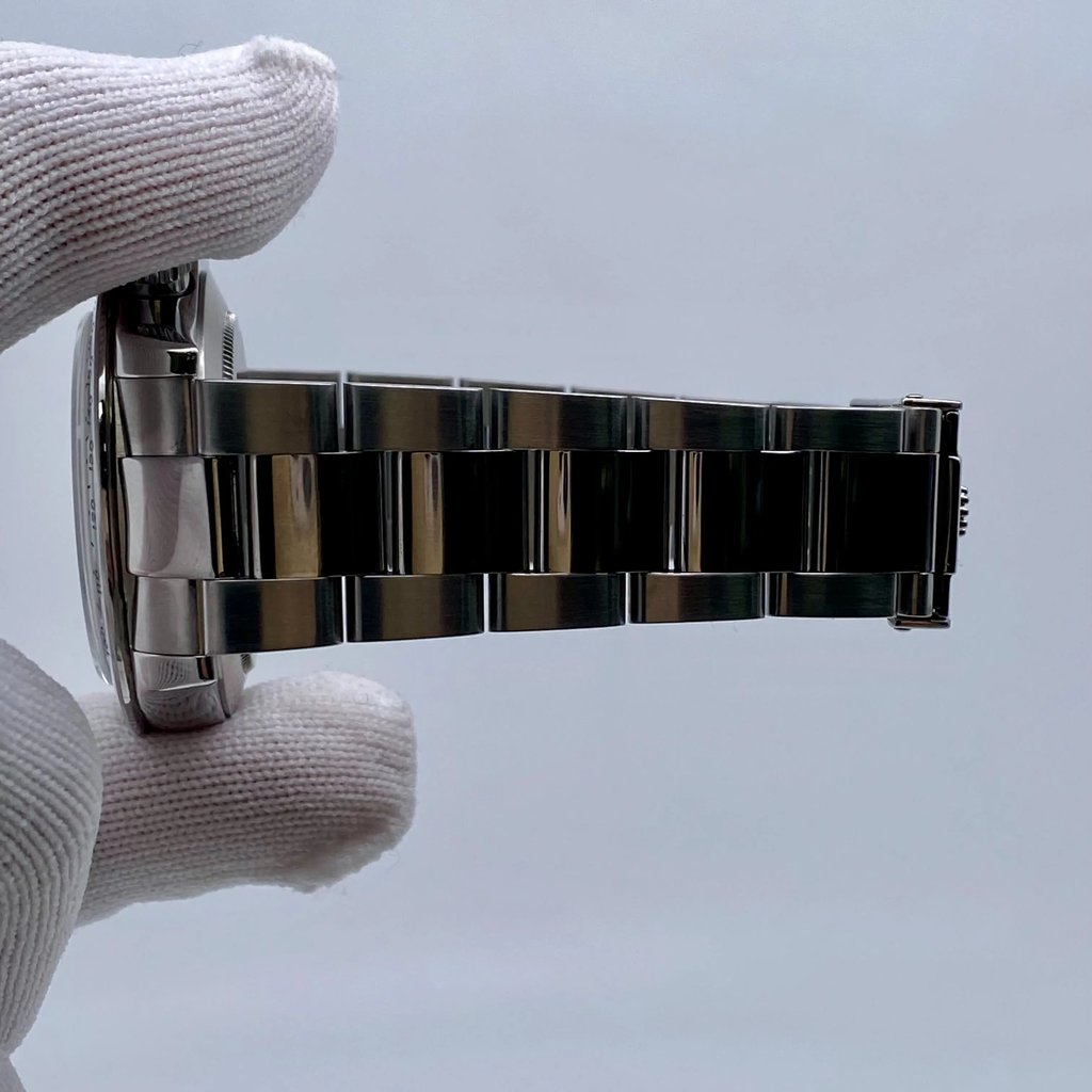 An image of a pre-owned men's Rolex wristwatch, featuring a stainless steel bracelet and case. The watch is held in a gloved hand, viewed from the side at a close distance, showing the bracelet links in detail. The chronograph and small seconds subdial features are not visible from this angle.