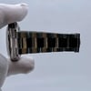 An image of a pre-owned men's Rolex wristwatch, featuring a stainless steel bracelet and case. The watch is held in a gloved hand, viewed from the side at a close distance, showing the bracelet links in detail. The chronograph and small seconds subdial features are not visible from this angle.