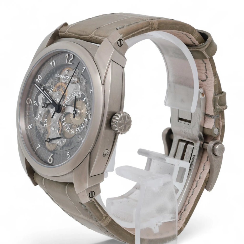 An image of a pre-owned Vacheron Constantin men's wristwatch displayed at a slight angle, showcasing the detailed watch face and luminous hands. The watch is positioned upright on a transparent stand, with its side and partially visible strap in view. The angle emphasizes the intricate design of the dial and the unique features of the timepiece, captured from a close distance.