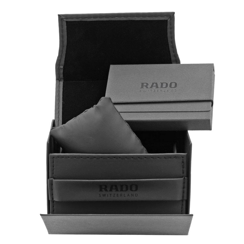An image of a wristwatch packaging set viewed from above. The image shows an open black box with the brand name "Rado" visible on the interior and exterior. Inside the box, there is a small rectangular booklet and a cushion, positioned centrally. The angle is straight-on and shot at a close distance, capturing the details of the box and its contents. The watch itself is not present in the image.