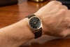 An image of a pre-owned men's Rolex wristwatch being worn on a wrist. The watch is viewed from a close-up, slightly angled perspective, showcasing its elegant black dial with rose gold accents and a brown leather strap. The background is a wooden surface, adding warmth to the overall composition.