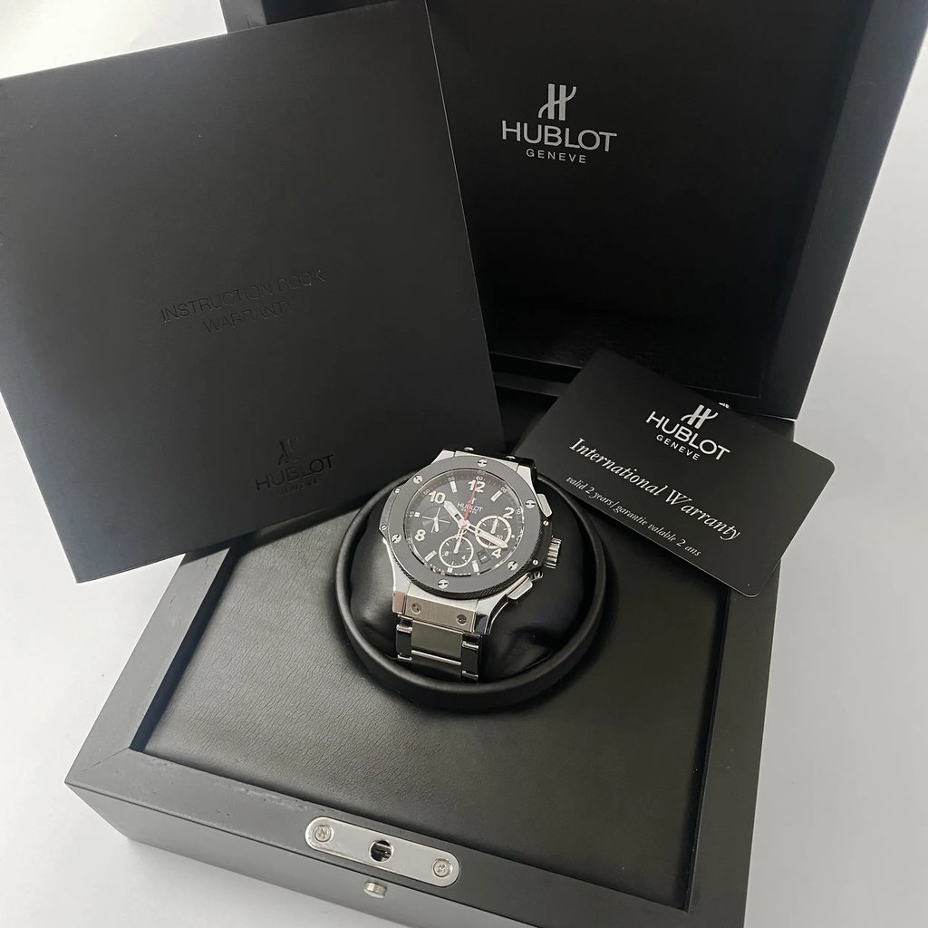 An image of a pre-owned wristwatch displayed prominently in the center, positioned on a cushioned holder inside an open box. The watch face is angled slightly upward, showing its detailed design. Surrounding the watch are branded documents, including an instruction booklet and an international warranty card, which are positioned to the left and right of the watch, respectively. The image captures the items from a close distance, allowing for a clear view of the watch and accompanying materials.