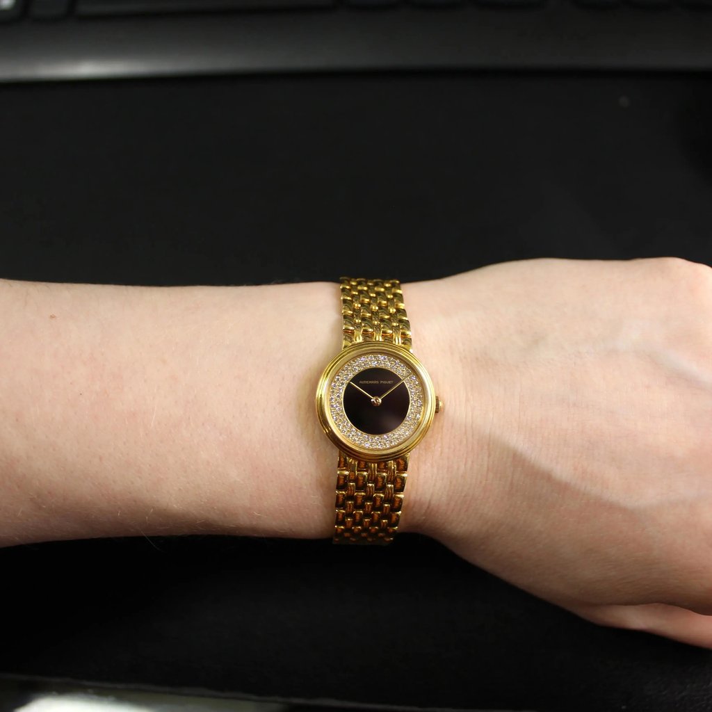An image of a pre-owned women's Audemars Piguet wristwatch on a wrist, viewed from above at a close range. The watch features a yellow gold bracelet and a round onyx dial with a diamond-studded bezel. The background is a dark surface with part of a keyboard visible at the top.