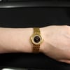 An image of a pre-owned women's Audemars Piguet wristwatch on a wrist, viewed from above at a close range. The watch features a yellow gold bracelet and a round onyx dial with a diamond-studded bezel. The background is a dark surface with part of a keyboard visible at the top.