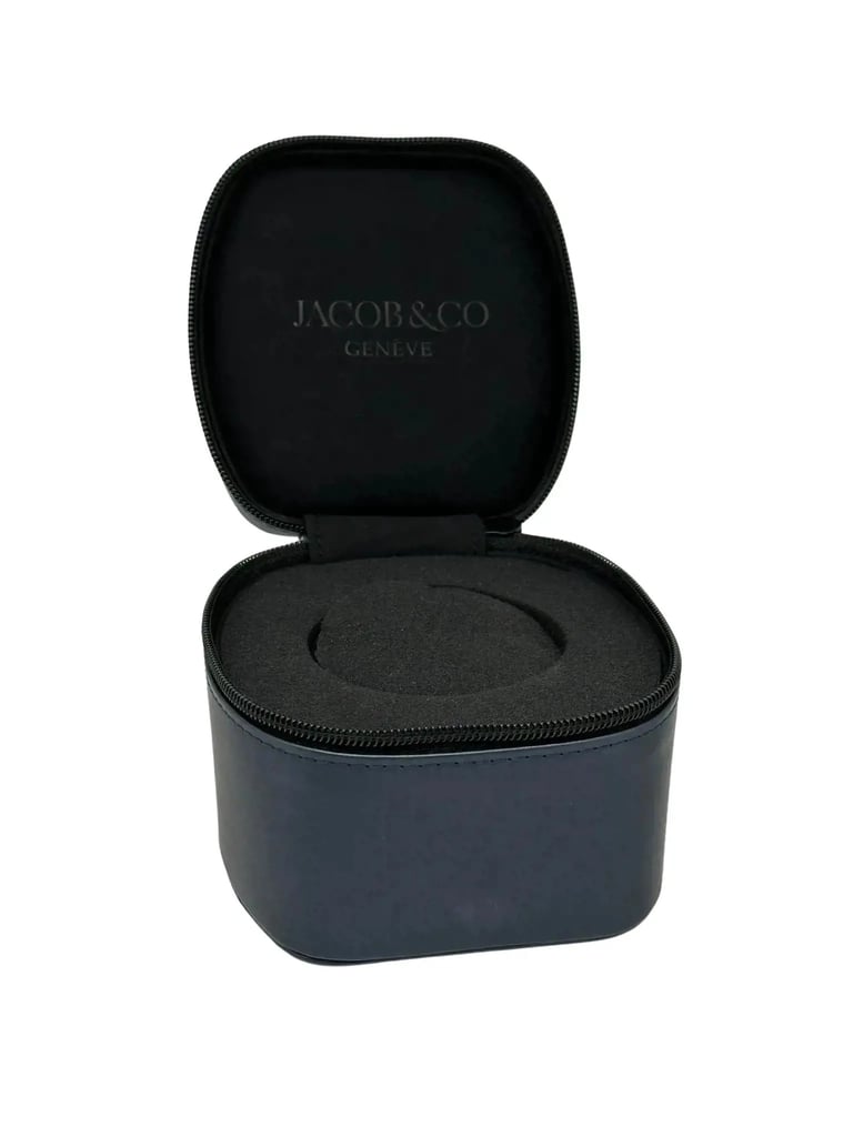 An image of a wristwatch box positioned centrally, viewed from a slightly elevated angle. The box is open, revealing a dark, cushioned interior designed to hold a watch. The brand "Jacob & Co Genève" is visible on the inside of the lid. The image is taken at a close distance, showcasing the box's texture and details.