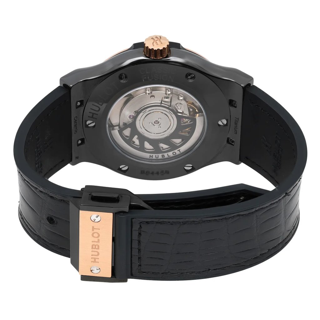 An image of a pre-owned men's Hublot wristwatch. The watch is positioned with the back facing the viewer, showcasing its intricate mechanical movement through a transparent case back. The angle is slightly elevated, providing a clear view of the watch's inner workings and branding. The distance is close, emphasizing details such as the gold-tone crown and clasp, as well as the black strap with a textured pattern.