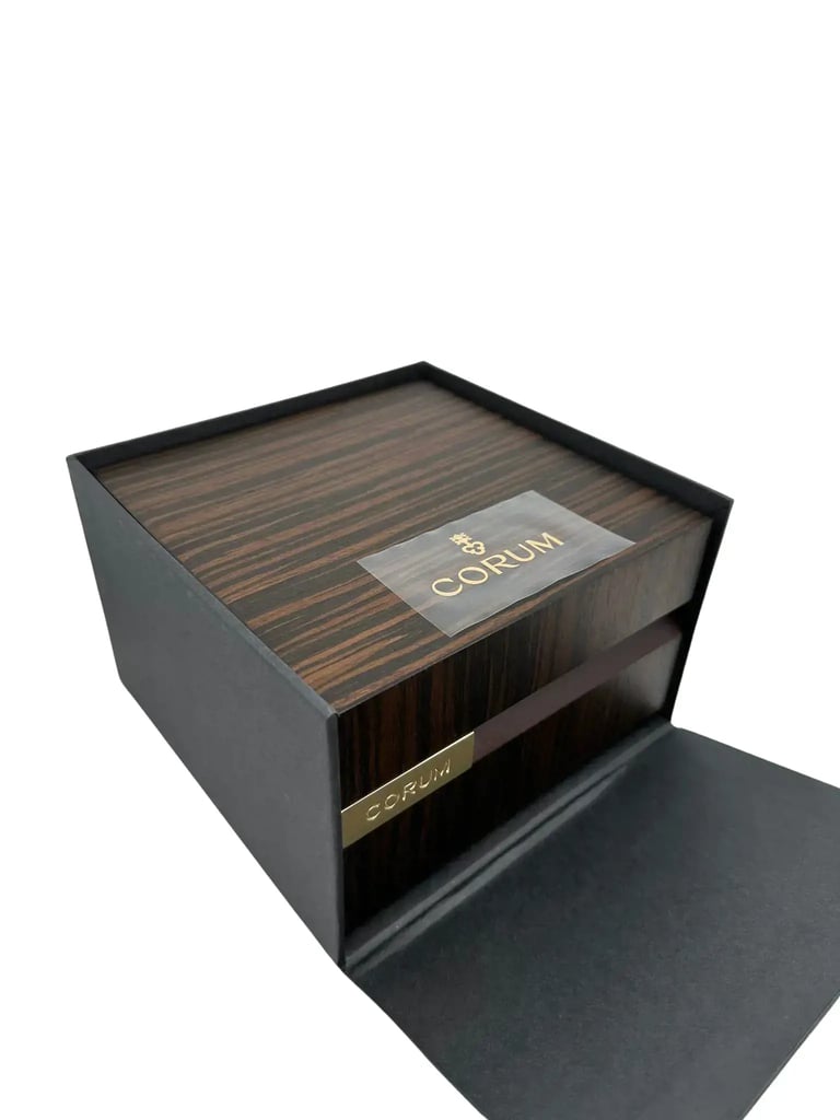 An image of a wristwatch box taken from a slightly elevated angle, showcasing the full view of the box and accompanying documents. The box is positioned centrally, with a dark wood finish and the brand name "Corum" prominently displayed in gold lettering on the top and front. The box is open, revealing an elegant presentation, but the wristwatch itself is not included in the image. The background is plain, emphasizing the box's details.