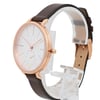 An image of a pre-owned Skagen wristwatch positioned at a slight angle, showcasing its round face and rose gold-tone bezel. The watch is displayed on a clear stand, with the face tilted slightly to the left. The white dial features minimalistic hour markers and a sub-dial with colorful markings. A brown leather strap is visible, with the buckle end in the background. The image is taken from a close distance, emphasizing the watch's elegant design details.