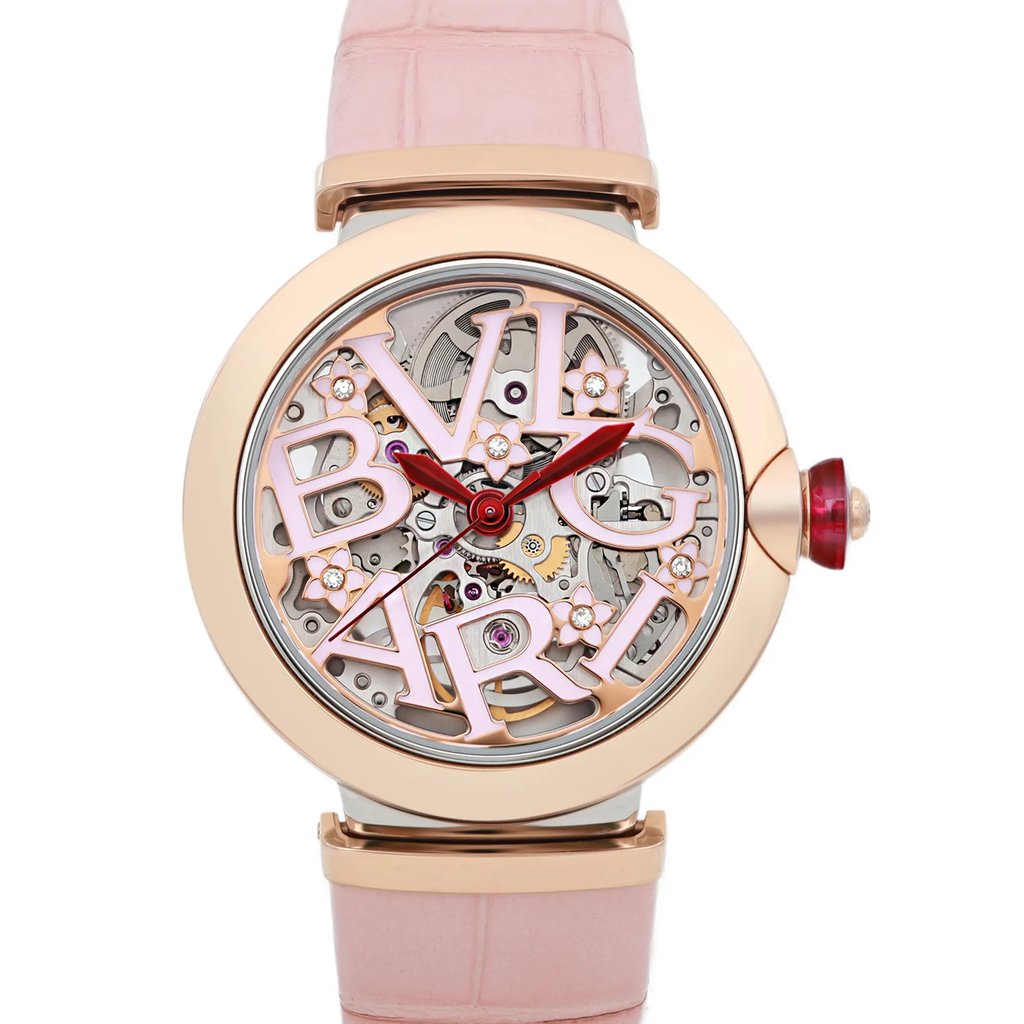An image of a pre-owned wristwatch taken from a front close-up angle, showcasing the intricate details of the watch face. The design features a rose gold-tone bezel with a visible mechanical interior and stylized lettering. The watch hands are red, and the strap is a soft pink color.