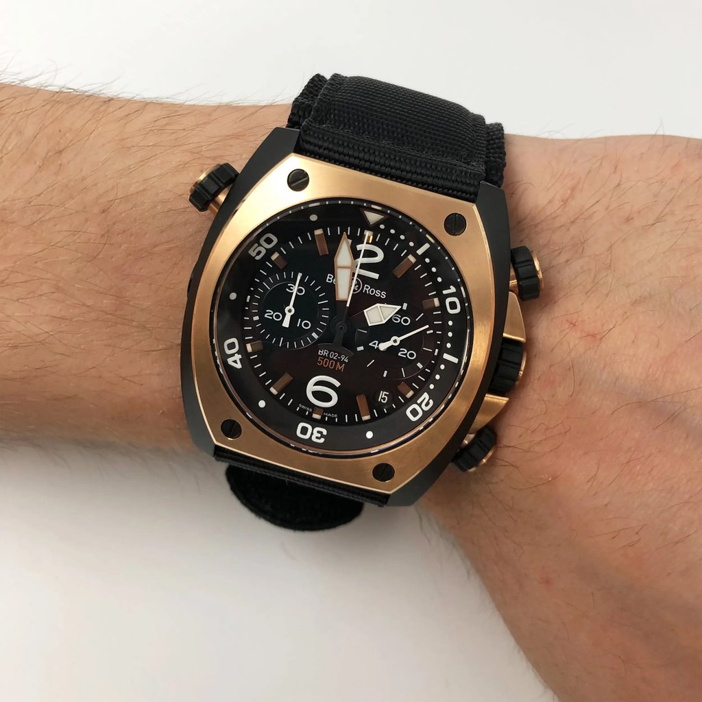 An image of a Bell & Ross men's luxury diver wristwatch, model displayed on a wrist, viewed from a front full frontal angle. The watch features a tonneau-shaped stainless steel case with a rose gold bezel, a black dial with large numerals, sticks indices, and a second hand for easy reading. It is attached to a black fabric/canvas band and the image is taken from a close distance, showing the watch in detail.
