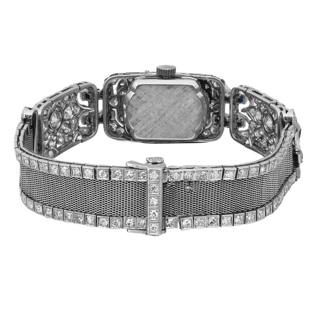 An image of a pre-owned Tissot women's wristwatch viewed from the back, showcasing the closed fold-over clasp. The wristwatch is positioned centrally and shot at a direct angle with a close-up distance, highlighting the intricate details. It features a rectangle-shaped case and band made of white gold, with a snap caseback. The band and case are adorned with diamonds, adding to its elegance.