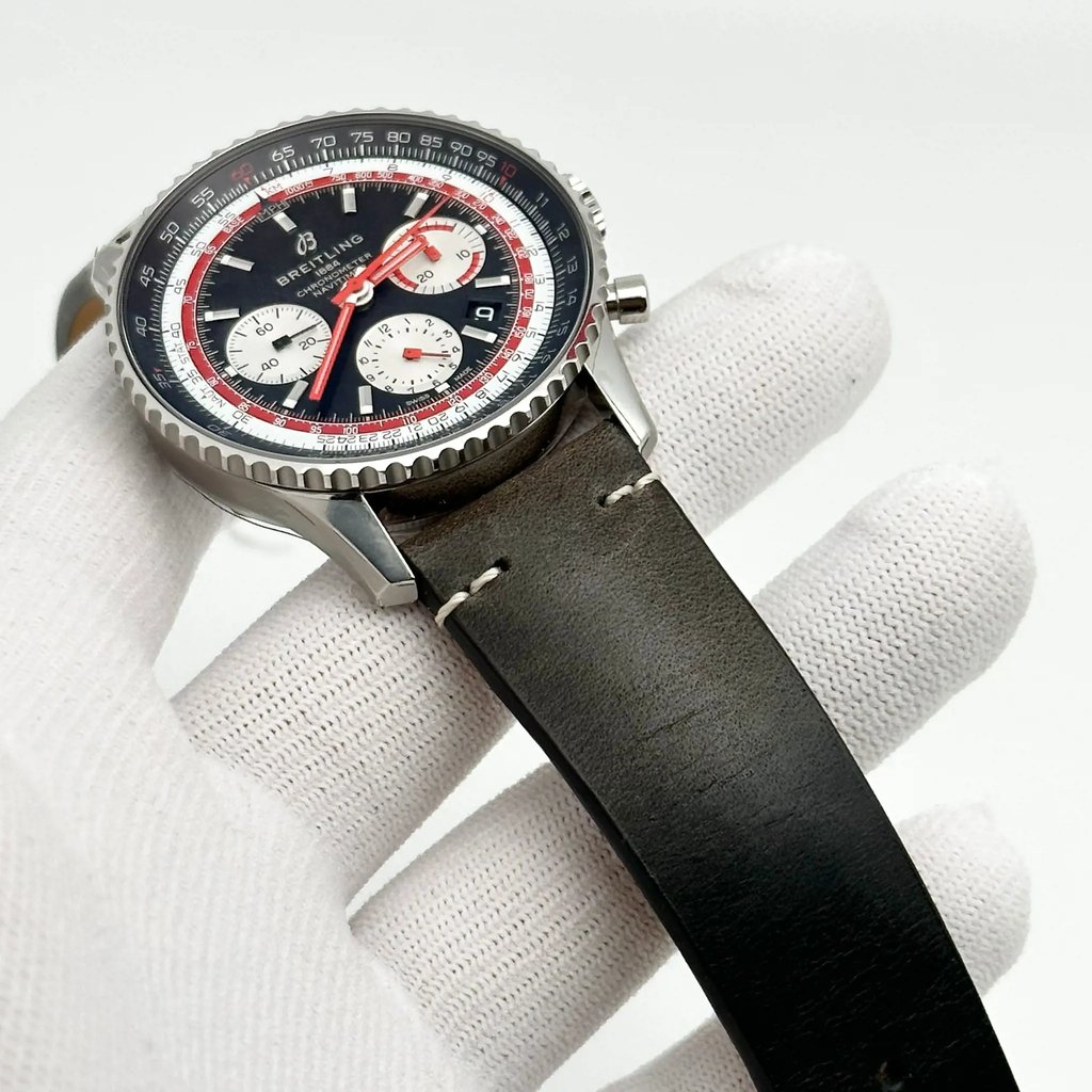 An image of a men's Breitling wristwatch, model displayed as a close-up at a three-quarter angle. The watch features a chronograph, date indicator, luminous hands, and a small seconds subdial. The timepiece, held by a gloved hand, shows a stainless steel case with a black and red dial, and is positioned against a white background. The black leather strap exhibits discoloration likely due to storage, suggesting the watch is a display model.