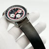An image of a men's Breitling wristwatch, model displayed as a close-up at a three-quarter angle. The watch features a chronograph, date indicator, luminous hands, and a small seconds subdial. The timepiece, held by a gloved hand, shows a stainless steel case with a black and red dial, and is positioned against a white background. The black leather strap exhibits discoloration likely due to storage, suggesting the watch is a display model.