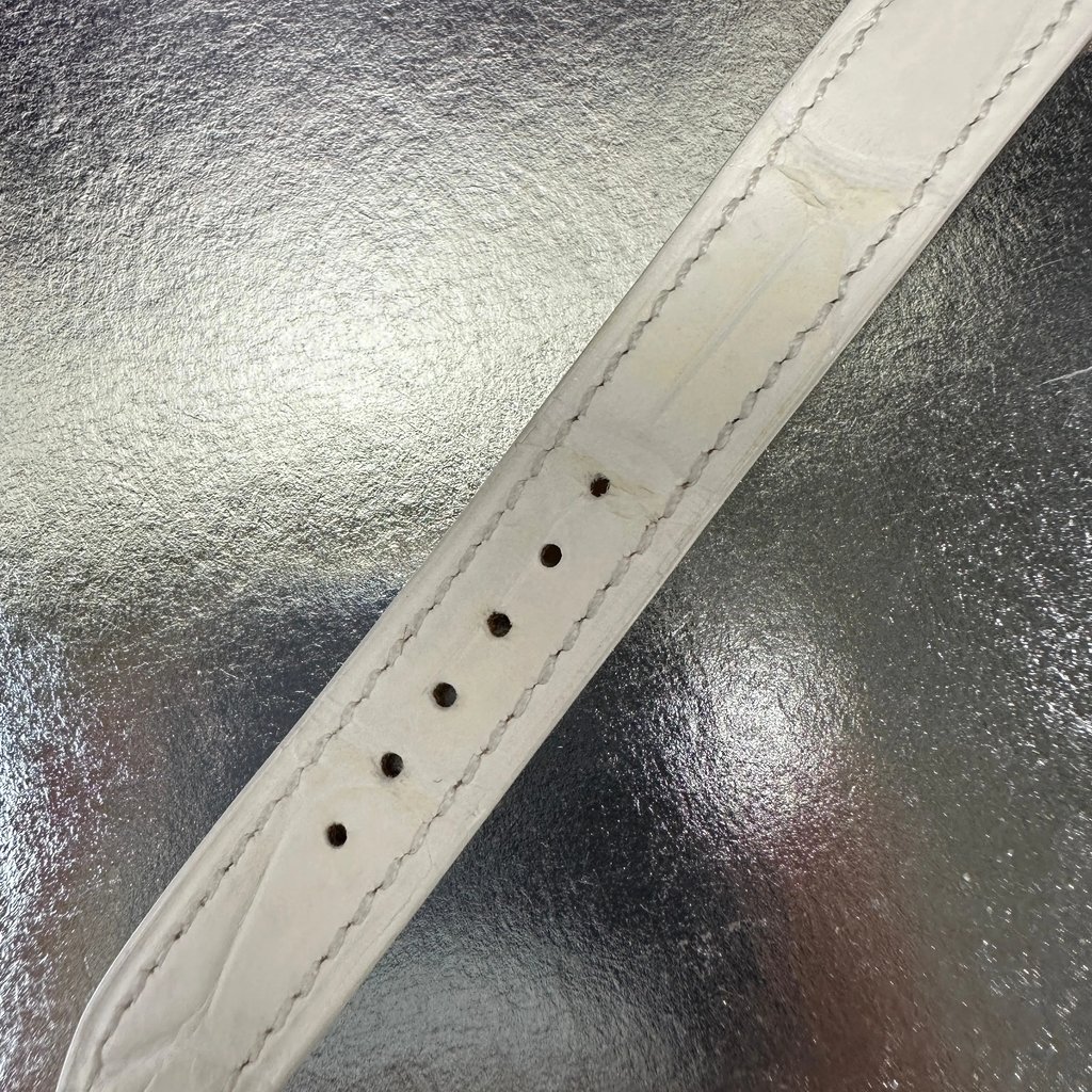 An image of a pre-owned Hermès women's wristwatch strap, viewed from a close-up angle. The strap is laid flat on a reflective surface, highlighting its light color and stitching details. The image captures the strap's adjustment holes and texture.